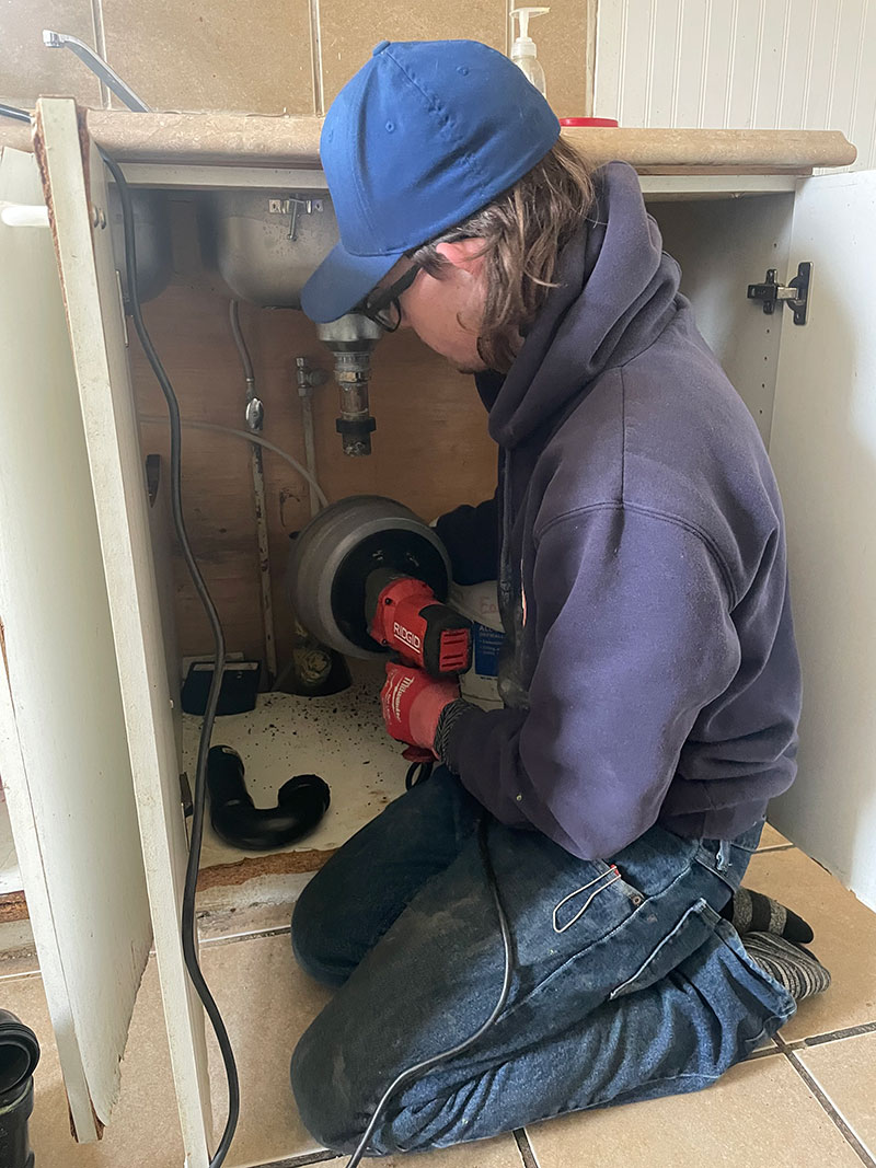A person kneeling under a sink uses a red power tool to work on plumbing pipes inside a cabinet, showcasing the effectiveness of Honey Badger Sewer solutions.