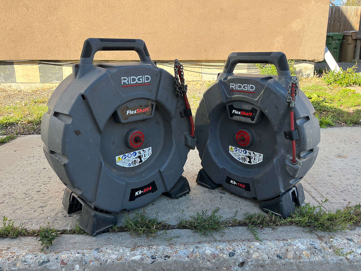 Two RIDGID FlexShaft machines (models K9-204 and K9-102) from Honey Badger Sewer are placed side by side on a concrete sidewalk next to a tan building.