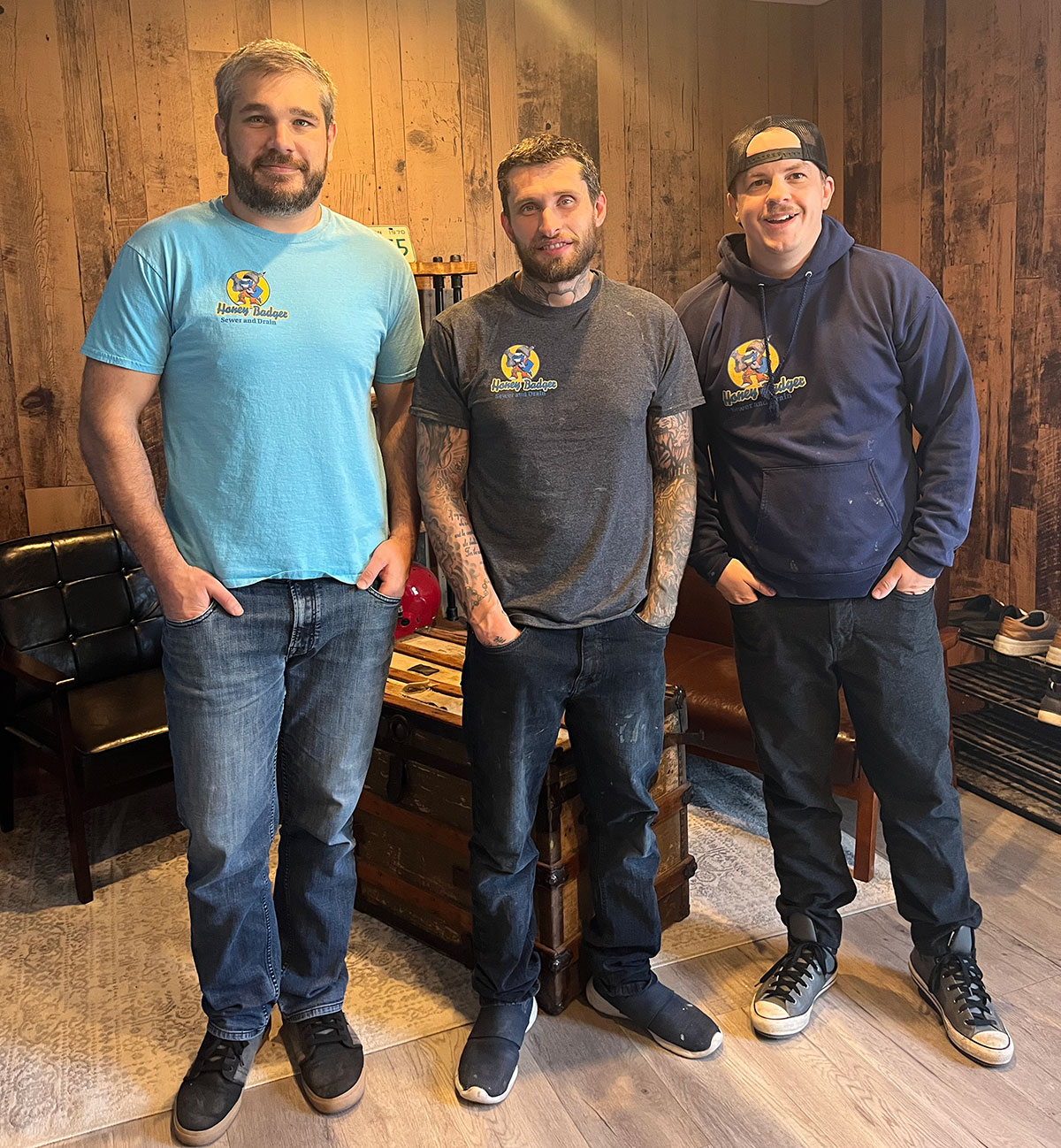 Three men stand indoors in front of a wooden wall, wearing casual clothes and shirts or hoodies with the Honey Badger Sewer logo.